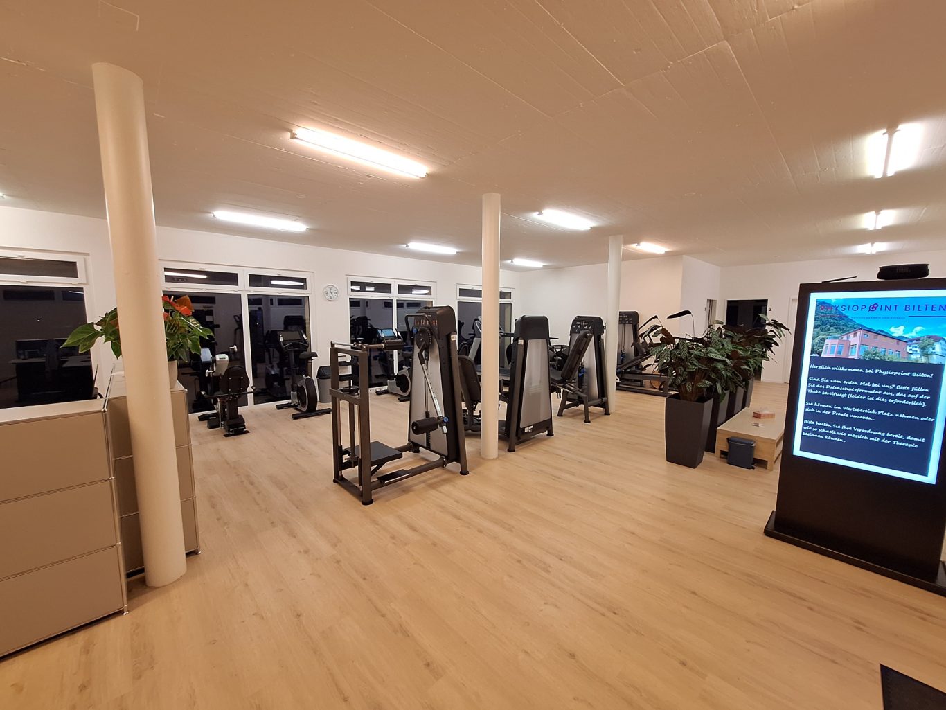 Moderne Fitnessstudio mit Trainingsgeräten und großen Fenstern, helle, freundliche Atmosphäre.